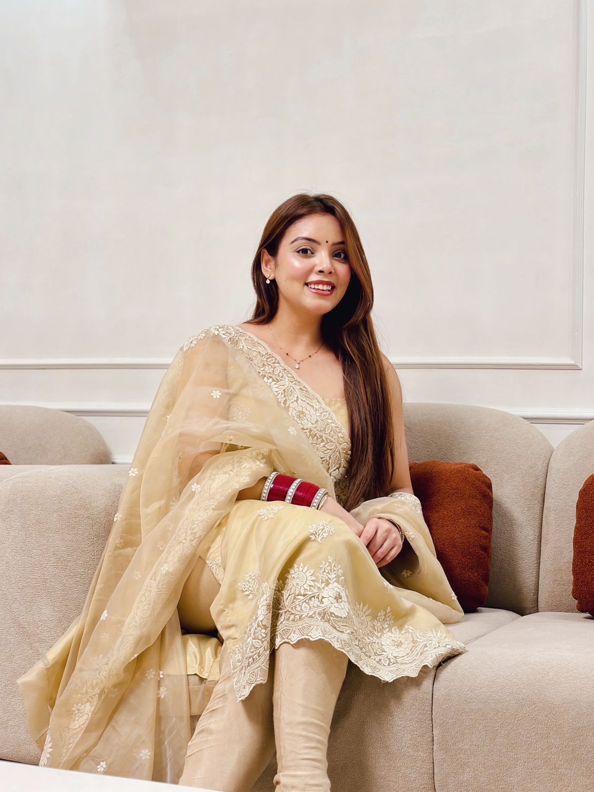 Sakshi Behl seated on an ivory sofa in an embroidered champagne suit
